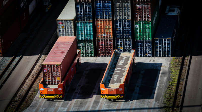 Two autonomous container transport vehicles operating in a shipping yard, surrounded by stacked shipping containers.