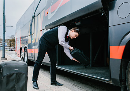 a driver or attendant, in a gray waistcoat, white shirt, and black trousers, loading a black suitcase into the luggage compartment of a large gray bu