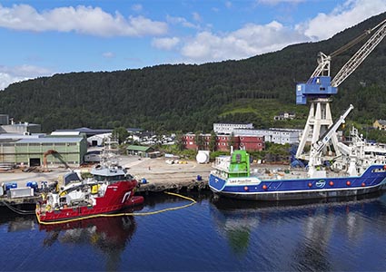 a shipyard scene in Norway, featuring specialized vessels docked at a industrial facility.