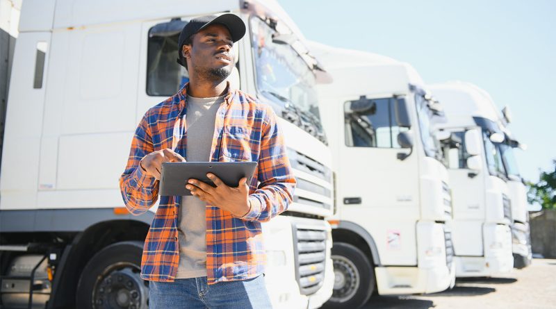 **Alt Text:** Man standing confidently in front of a fleet of commercial freight trucks at a logistics depot in the United States.