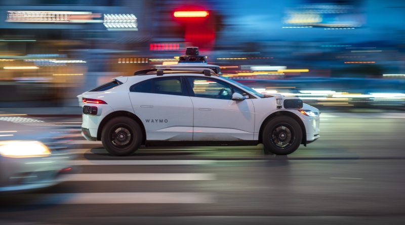 A Waymo autonomous vehicle driving through a busy urban street, representing the launch of the Waymo Premier subscription service and the evolution of robotaxi business models.