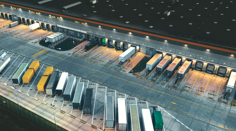 Aerial view of a modern logistics warehouse with multiple loading bays, trailers and freight trucks positioned for distribution, illustrating large-scale warehousing and supply chain operations.