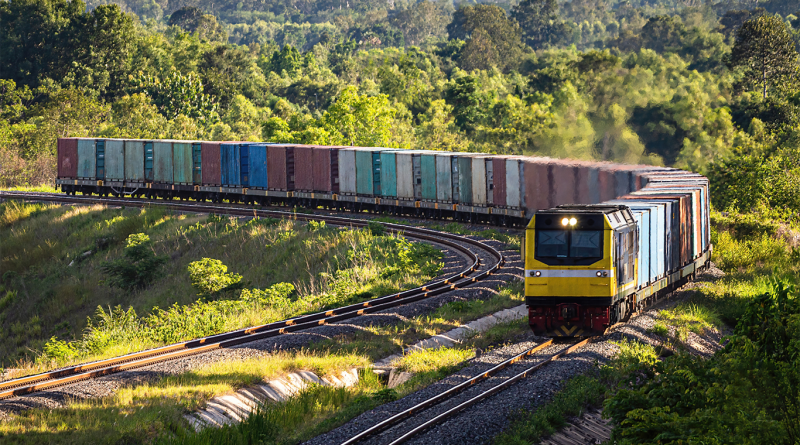 Freight train carrying shipping containers along curved railway tracks through a green forest landscape.