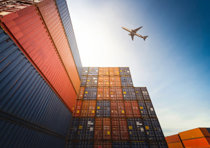 a large stack of colorful freight shipping containers with a cargo airplane flying overhead against a bright sky