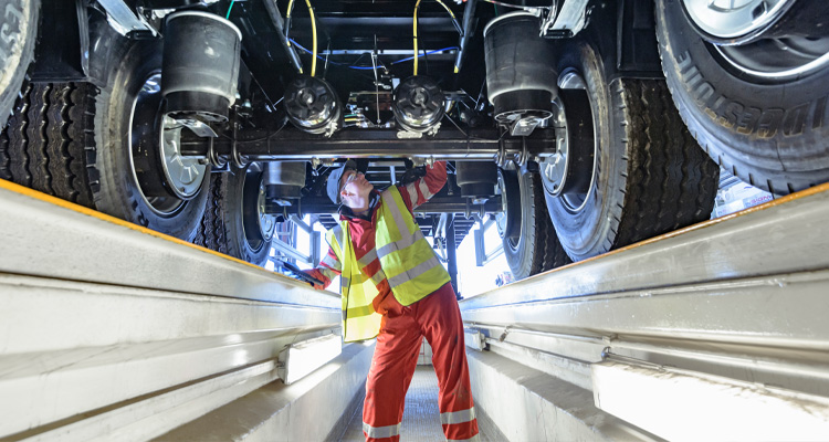 A mechanic inspects the undercarriage of a heavy-duty commercial vehicle or trailer from an inspection pit in a workshop