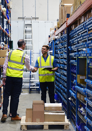 two warehouse workers shaking hands while reviewing stock near shelves and a pallet of boxes